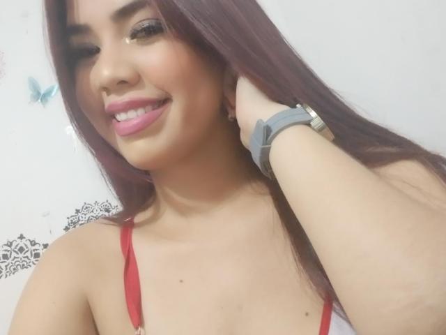 AngelVeriiHot with long brown hair, pink lipstick, and red bra, smiling with one eye closed, touching her head with a gray wristwatch, showing her tits and cute face.