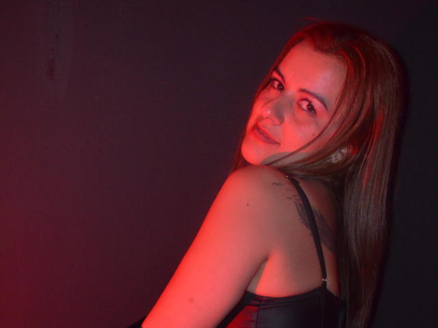 PerlaDivaio with long brown hair, looking over her shoulder, smiling, wearing a black leather top, red light on her right shoulder, subtle tattoo, soft skin, sexy vibe.