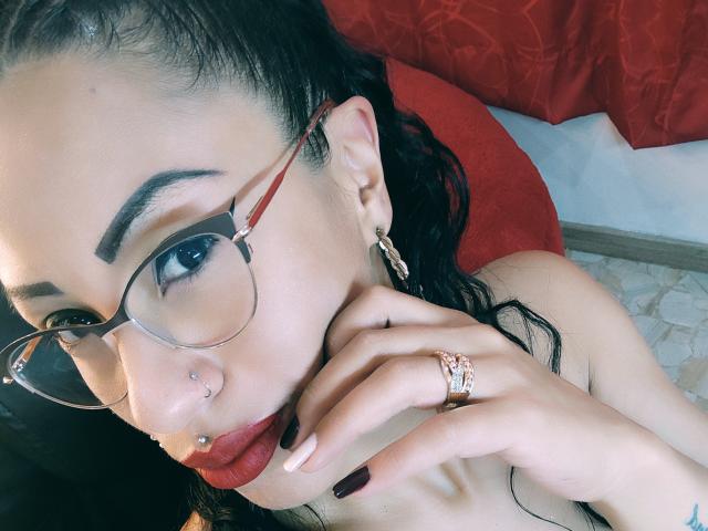 LunaCandy con gafas, lápiz labial rojo, piercing en la nariz y anillo, cabello negro, aretes y uñas cuidadas, tocándose los labios, usando un anillo rosa, con una mirada sexy y segura..