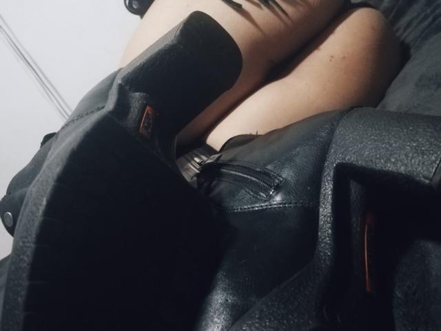 Le gambe di LeanneFoxy, con stivali di pelle nera, cosce scoperte, pelle morbida, ginocchia leggermente piegate, texture della pelle, suole degli stivali a fuoco, il suo sedere appena visibile, angolazione intima, ombre delicate..