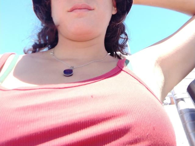 Semda with pale skin, wavy brown hair, wearing a red ribbed tank top, showing her tits, purple pendant necklace, arm raised, blue sky background, sunlight.