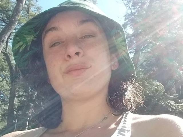 Semda, con un gorro verde, cabello ondulado castaño, camiseta blanca y una sonrisa sutil, con la luz del sol brillando, mostrando una apariencia relajada y natural, con un collar pequeño..
