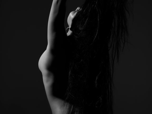 ArielleWilde with long black hair, arms raised, exposing her tits, and a slim, toned, dark-skinned body in a black-and-white, side-profile shot.