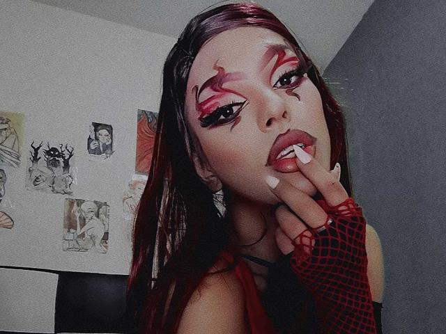 LucyVendeta, con cabello rojo oscuro, maquillaje rosa y negro intenso, y guantes de malla roja, mordiéndose el dedo con uñas blancas, mostrando sus labios rojos y carnosos y sus dientes blancos..