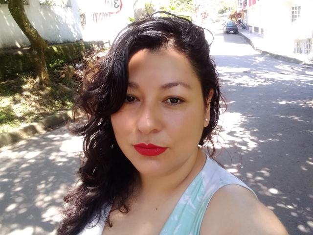 SakotoDika with wavy black hair, red lipstick, and a light blue shirt, standing on a sunlit street, looking at the camera with a slight smile.