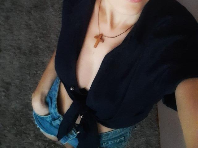 AnnieXCute's black shirt tied below her chest, showing cleavage, wearing a cross necklace and blue denim shorts, with hands in her shorts, on a grey carpet.