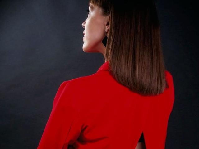 AnnieXCute with straight brown bob, wearing a red jacket, facing left, showing her profile with subtle makeup, dark background.