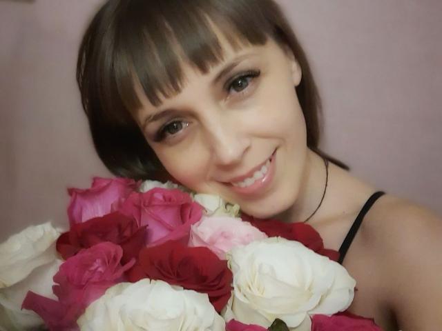 AnnieXCute with short brown hair and bangs, smiling, holding a bouquet of red, pink, and white roses, wearing a black strap top, showing her cute face and teeth.