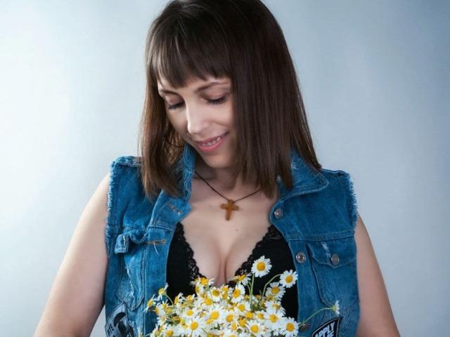 AnnieXCute with brown bob, blue denim vest, black lace bra, holding yellow daisies, wearing cross necklace, looking down, smiling, showing her tits, in a relaxed pose.
