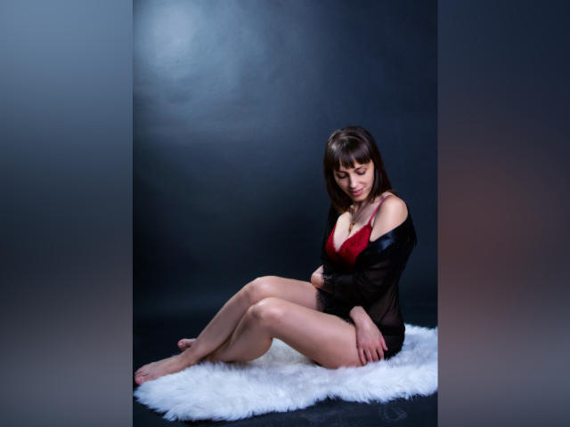 AnnieXCute sits on a white furry rug, wearing a red bra and black sheer robe, with her tits visible, legs crossed, and arms resting on her knees.