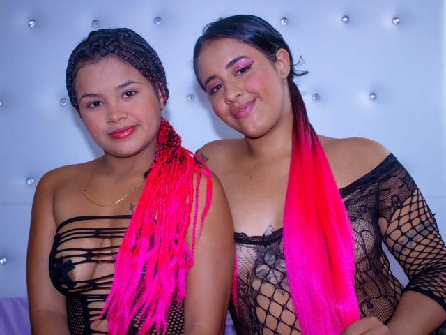 EvaAndLisbeth avec des tresses et des couettes roses, EvaAndLisbeth avec une robe en maille noire et un foulard rose, toutes deux montrant leurs seins à travers la maille, souriantes. Peau claire, latina, sexy..