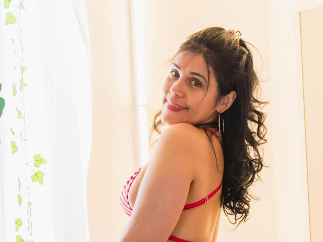 Girasol Walton, avec ses cheveux noirs ondulés, son soutien-gorge rouge et ses boucles d'oreilles, sourit à l'appareil photo, montrant sa poitrine, et se tient près d'une fenêtre avec des rideaux blancs..