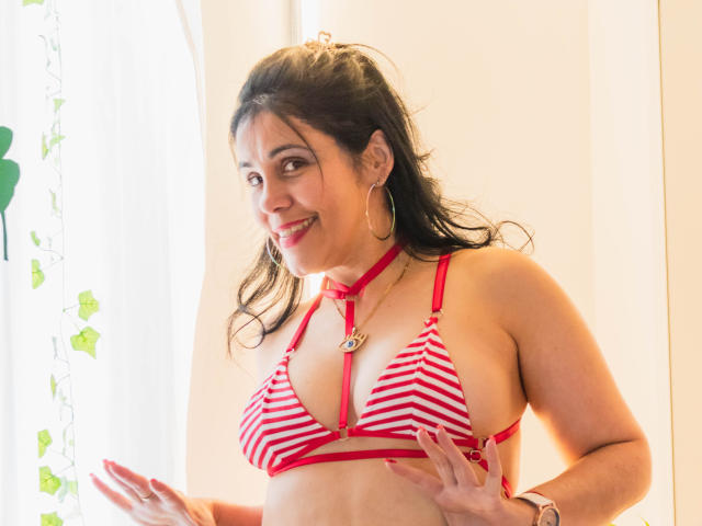 GirasolWalton, a curvy woman with medium brown skin, wearing a red and white striped bra with straps, smiling at the camera, has small boobs, hoop earrings, and a necklace.