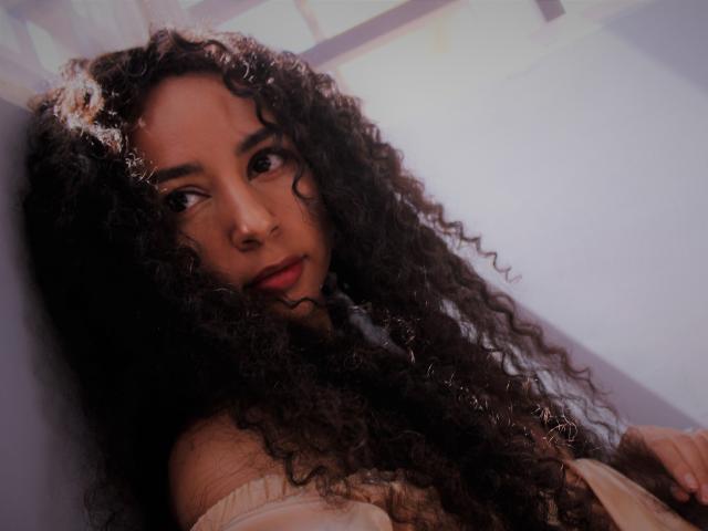 SexyKonnyX with curly dark brown hair, red lipstick, and off-shoulder beige top, leaning against a wall, looking slightly to the side with a subtle, sexy smile.