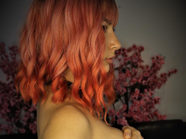 SexyKonnyX met rood, golvend haar, in profiel, die haar kleine, onbedekte borsten laat zien met een zwarte choker, op de achtergrond roze kersenbloesems, met haar blik naar beneden gericht, met zachte belichting..