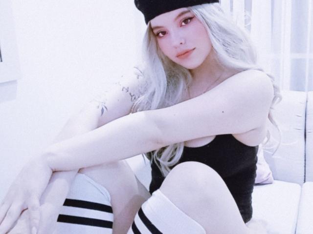 ElyMoon, avec ses longs cheveux blancs, portant un bonnet noir et un débardeur, assise les genoux repliés, montrant des chaussettes blanches à rayures noires, et un tatouage sur son bras..