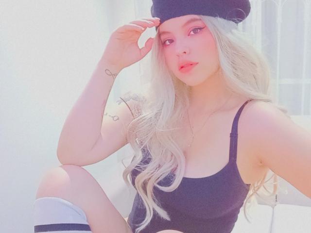 ElyMoon tiene el pelo rubio, largo y ondulado, y lleva una camiseta negra y un gorro, con maquillaje rosa, un pequeño tatuaje de corazón y una media blanca que le llega a la rodilla, mirando a la cámara..
