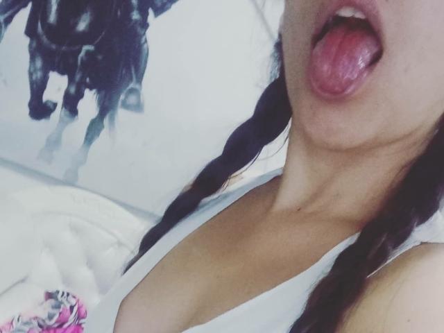 SammyJuts, Zunge heraus, weißes Tanktop, sichtbarer Ausschnitt, geflochtenes braunes Haar, helle Haut, weicher Fokus, rosa Blumendecke unten links, verschwommenes schwarzes Pferd an der Wand..