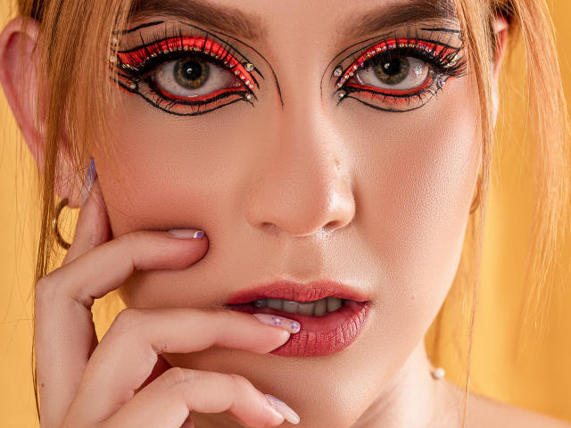 MelisaLee's face, red and black eyeliner, blue eyes, light skin, pink lips, blonde hair, fingers touching her lip, yellow background, glossy nails, subtle makeup, seductive expression.
