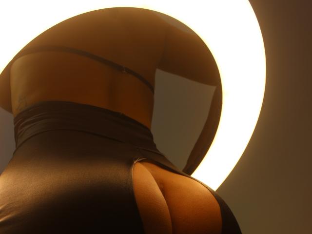 Il sedere di SexyKonnyX, in leggings neri aderenti, mostra un'ampia apertura che rivela la pelle abbronzata e una parte della sua schiena. Illuminato da una luce circolare proveniente dall'alto..
