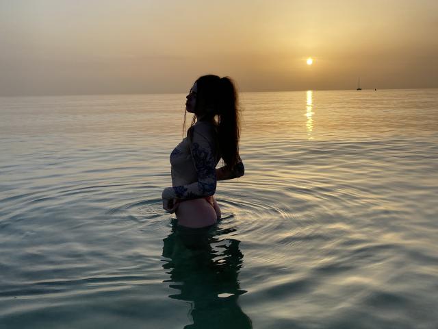 ValeriMair staat in het water, silhouet tegen de zonsondergang, met een top met veters, die haar borsten en billen laat zien, lang haar, zonnebril en een roze string. Er ontstaan rimpelingen rondom haar..