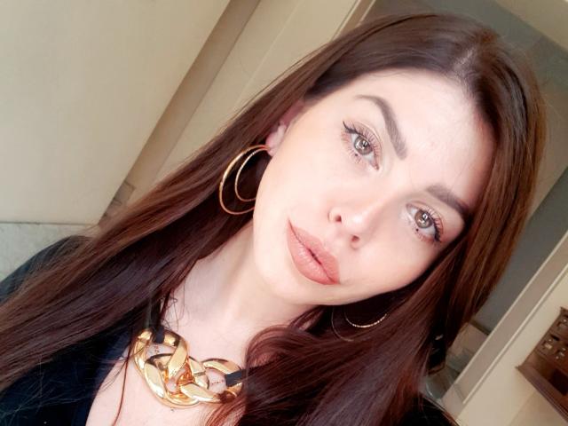 ValeriMair with long brown hair, wearing gold hoop earrings and a chunky gold necklace, has blue eyes and pink lipstick, looking slightly to the right.