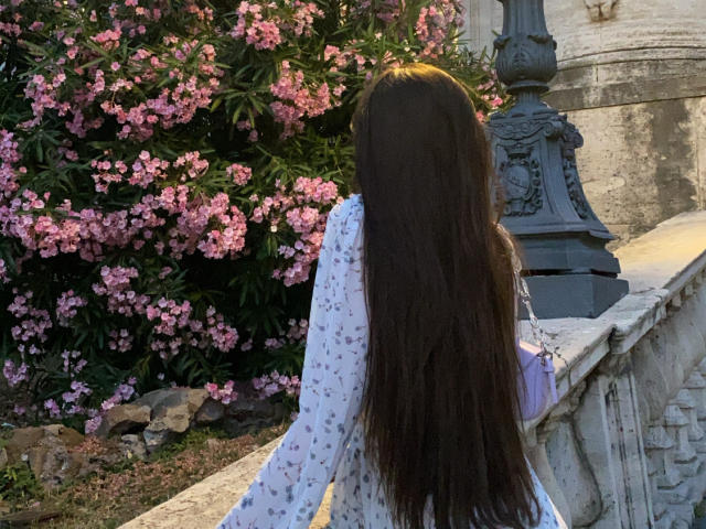 ValeriMair with long brown hair, back to camera, wearing a white floral dress, standing next to a stone pillar with pink flowers in the background.