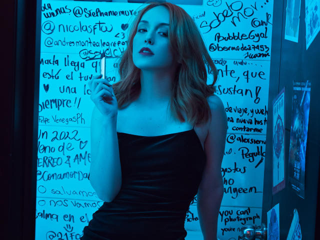 MollyMarmalade with red hair, wearing a black strapless dress, standing with a cigarette, blue-tinged lighting, looking confident, showing off her tits, and a slight smile.