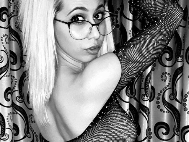DiamondHottieX met platinablond haar, een bril en glinsterende mesh-mouwtjes, kijkt om en heeft een fronsende uitdrukking, waarbij haar blote rug en schouders te zien zijn. Zwart-wit afbeelding..