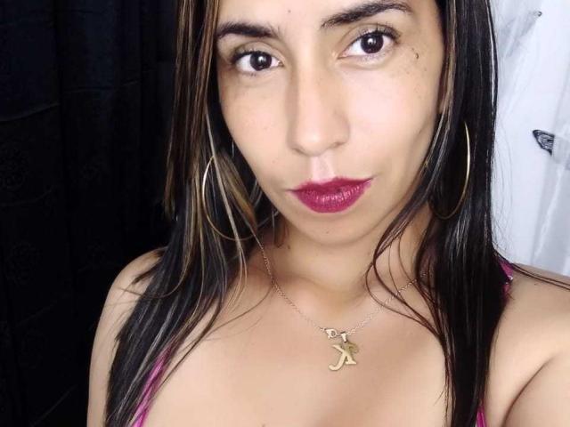 KatyKatt with medium brown skin, dark brown eyes, and straight black hair wearing pink lingerie, gold hoop earrings, and a Y necklace, has glossy red lipstick and a pout.