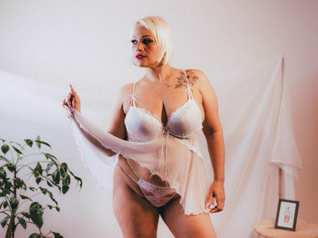 GianellaCox with blonde bob, wearing white lace lingerie, holding sheer fabric, exposing her tits and pussy, tattoo on left shoulder, standing near green plant, soft pink lighting.