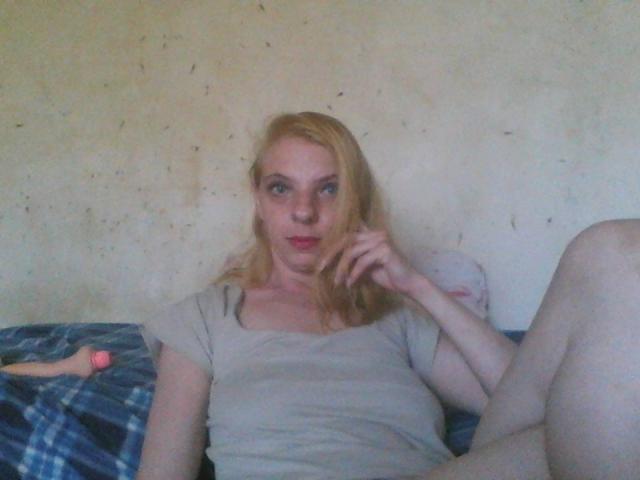 BlondyMarianne tem pele clara, cabelo loiro, olhos azuis e seios pequenos. Ela está vestindo uma camiseta cinza, sentada com as pernas afastadas, usando um relógio rosa, em uma cama azul xadrez..