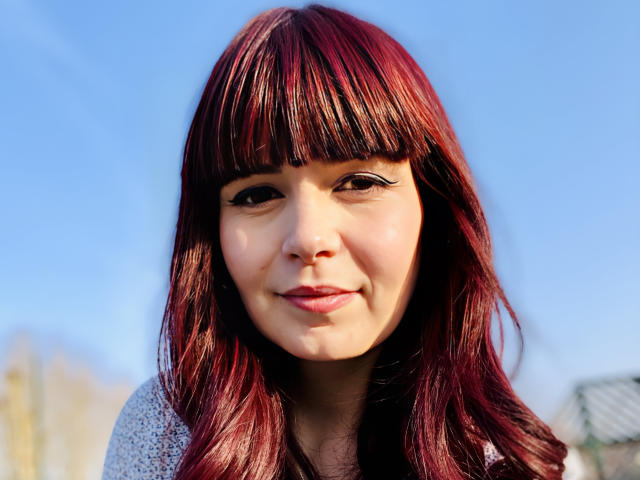 BellaHottest has shiny red hair, bangs, and black eyeliner, with a cute smile, wearing a gray sweater, showing her face and upper chest. Bright blue sky.