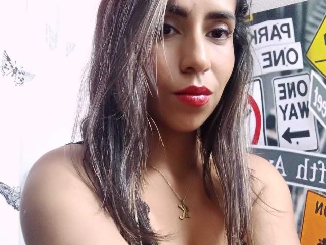 KatyKatt has medium brown skin, long dark brown hair with blonde highlights, red lipstick, and a gold necklace with an "L" pendant. She's looking at the camera with a slight smile.