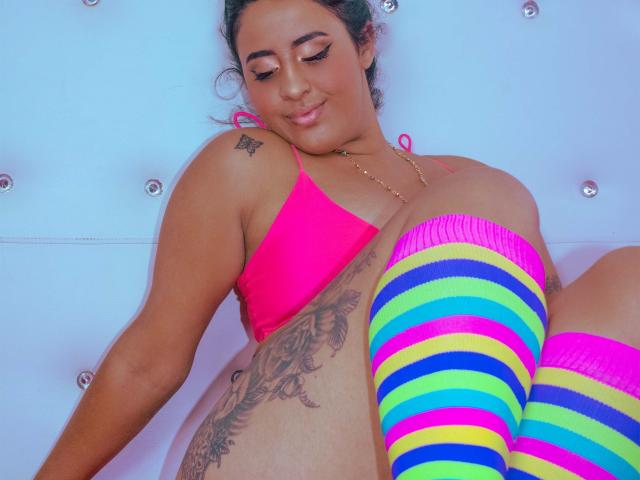 EvaAndLisbeth, avec une peau bronzée, un haut de maillot de bain rose, des bas rayés colorés, de grandes seins, un tatouage sur le côté, des cheveux verts, souriant, assise, fond bleu. Tatouage de seins, tatouage de fesses..