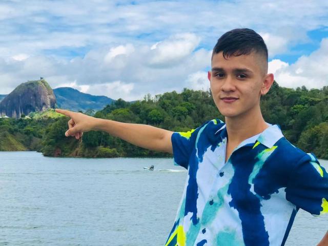 LoganD, short dark hair, wearing a blue and white abstract shirt, points to the left, smiling, standing by a water body, with a rocky hill and trees in the background.
