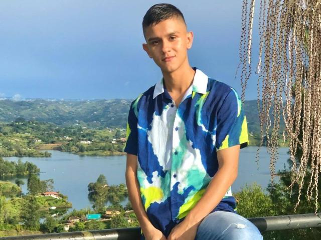 LoganD with short brown hair, wearing a blue, green, and white tie-dye shirt, and ripped jeans, sitting on a balcony with a hanging plant, looking at the camera.