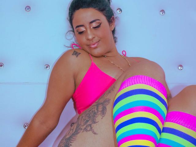 EvaAndLisbeth con reggiseno rosa, calzini colorati a righe, tatuaggio sul fianco, capelli scuri raccolti in uno chignon, sorridente, che guarda in basso, carina, formosa, pelle abbronzata, piccola collana d'oro, sfondo di parete bianca..