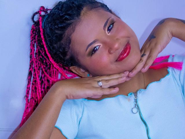 Eva y Lisbeth, con cabello trenzado rosa, lápiz labial rojo, sonriendo, con las manos cerca del rostro, vistiendo una blusa azul claro, un anillo y un collar, sobre un fondo blanco..