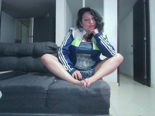 SakotoDika, sitting on a gray sofa with legs spread, wearing a blue and green tracksuit, hand on chin, looking at camera, bare feet, black hair, red lipstick.