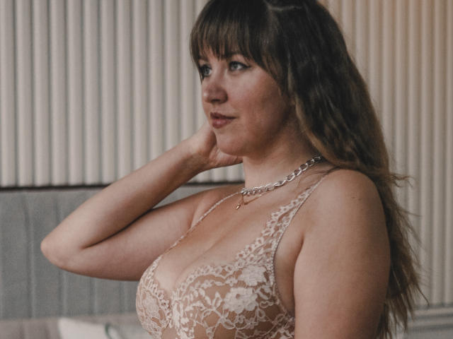 Jessica Logan mit braunen Haaren, trägt ein Spitzen-Bralette, zeigt ihre Brüste, eine Kette um den Hals, Hand am Wangenknochen, Seitenansicht, blickt in die Kamera, sanftes Lächeln, Halbnahaufnahme, Innenaufnahme..