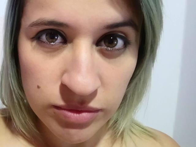 Le visage de BijouBlonde69, avec ses cheveux blond-vert, ses yeux marron et une petite tache de naissance sur sa joue gauche, affiche une expression neutre, et ses lèvres roses sont légèrement entrouvertes..