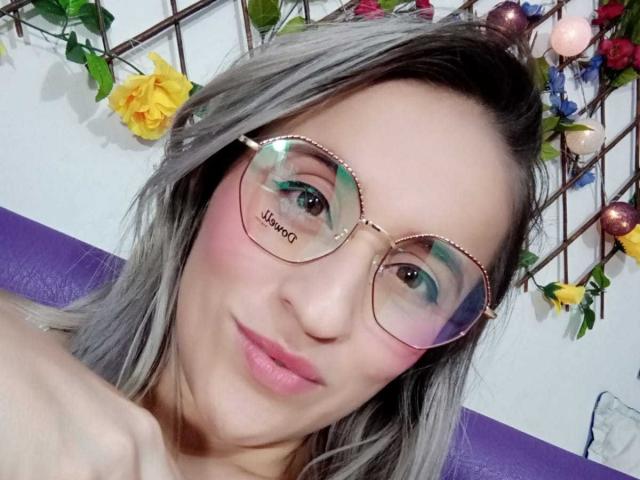 BijouBlonde69 tiene rubor rosado, sombra de ojos verde azulada, lápiz labial rosa y usa gafas reflectantes de color rosa dorado con la palabra "jumano" en la lente izquierda, sonriendo y con cabello gris..