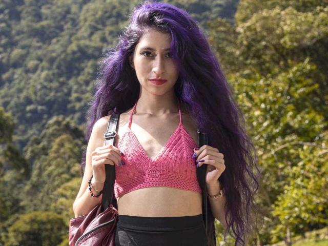 Julia mit langen, purpurfarbenen Haaren, einem pinken, gehäkelten Top, kleinen Brüsten, die eine schwarze Rucksack hält, einen schwarzen Rock trägt, draußen steht, im Hintergrund ein Wald zu sehen ist, wirkt ernst und hat die Hände an den Trägern des Rucksacks..