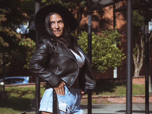 AmelieBlake with dark leather jacket, white tee, ripped blue jeans, black hat, hand on hip, smiling, medium tits, brown skin, long black hair, standing outdoors, sunlight, casual.
