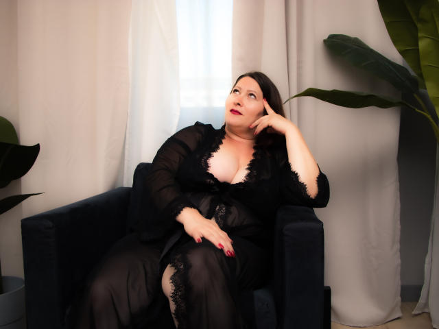 SoniaLaBelle, uma mulher corpulenta com seios médios, está sentada em uma cadeira preta, vestindo um vestido preto de renda transparente, com unhas vermelhas, cabelo escuro e batom vermelho, olhando para cima..