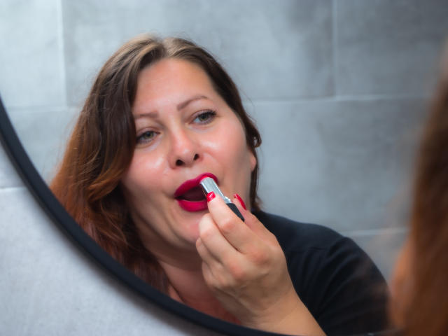 SoniaLaBelle with red lipstick, applying more with a red lipstick tube in front of a mirror, showing her slightly open mouth and focused expression.