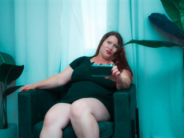 SoniaLaBelle has big boobs and a curvy ass, sitting in a green chair, holding a phone, with long brown hair and red lipstick, wearing a black dress.