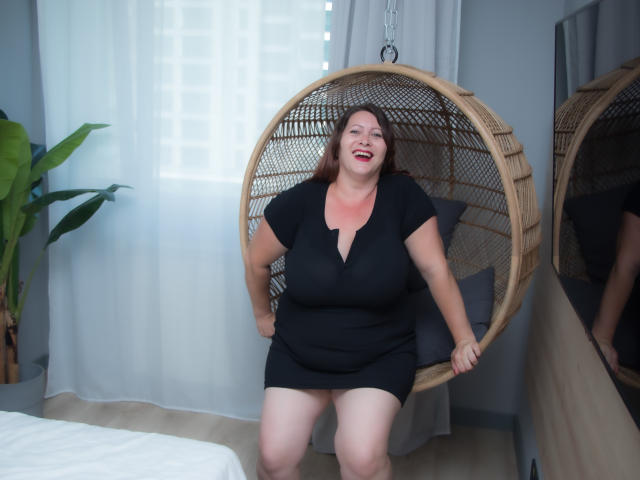 SoniaLaBelle, plus-sized with pale skin and red hair, sits in a wicker hanging chair, wearing a black dress, smiling, hands on chair, reflection in mirror, plant in corner.