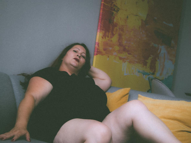 SoniaLaBelle, plus-sized with pale skin, black dress, and brown hair, lounges on a grey couch, legs spread, yellow pillow to her right, painting with red and yellow brushstrokes.